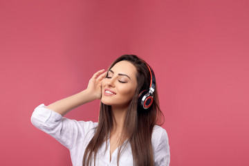 Fototapeta premium Beautiful woman smiling with smartphone in hands enjoying favorite music, via wireless headphones over pink background. Girl uses wireless earphones and dancing..