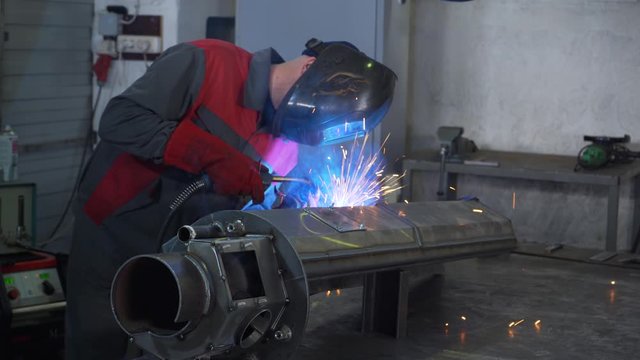 Professional welder is finishing work on complex metal device, welding energy, blue flame, sparks and smoke, man in protective suit, welding mask, tight gloves, factory floor, welding machine, gas