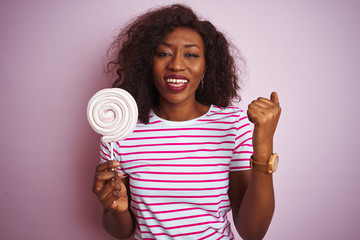 Young african american woman eating sweet candy standing over isolated pink background screaming proud and celebrating victory and success very excited, cheering emotion