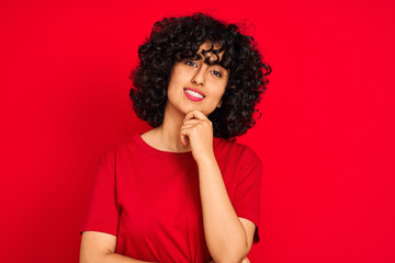 Fototapeta na wymiar Young arab woman with curly hair wearing casual t-shirt over isolated red background looking confident at the camera smiling with crossed arms and hand raised on chin. Thinking positive.