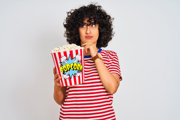 Young arab woman with curly hair holding pack of popcorns over isolated white background serious face thinking about question, very confused idea