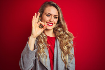 Young beautiful business woman wearing elegant jacket standing over red isolated background smiling positive doing ok sign with hand and fingers. Successful expression.