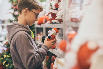 Naklejka premium Young boy looking a puppet of Santa Claus, thinking about beautiful gifts. Kid chooses christmas decoration for his home in a shop full of beautiful adorns. Holidays, party and new year eve concept.