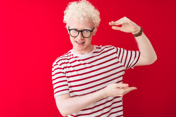 Young albino blond man wearing striped t-shirt and glasses over isolated red background gesturing with hands showing big and large size sign, measure symbol. Smiling looking at the camera. Measuring