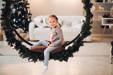 Stock photo portrait of little girl sitting on soft pillow of circle swing decorated with Christmas fir tree and pine cones. She is smiling at camera. Cozy sofa and Christmas tree in background.