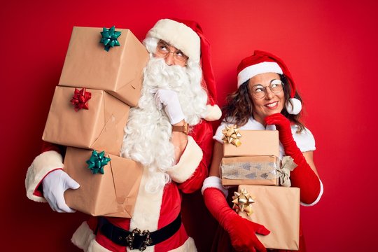 Middle age couple wearing Santa costume holding tower of gifts over isolated red background with hand on chin thinking about question, pensive expression. Smiling and thoughtful face. Doubt concept.