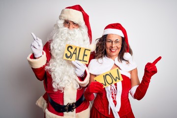 Couple wearing Santa costume holding wow and sale banner over isolated white background with a big smile on face, pointing with hand finger to the side looking at the camera.