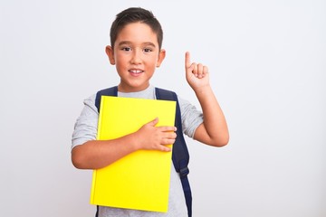 Beautiful student kid boy wearing backpack holding book over isolated white background surprised...