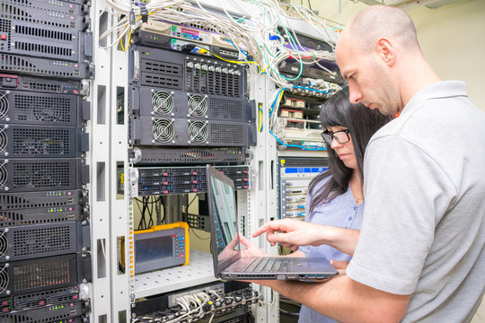 A Man And A Woman With A Notebook Work In A Server Room. Experts Look At The Computer Screen In The Data Center. System Engineers Customize Hardware.