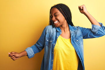Young african american woman wearing denim shirt standing over isolated yellow background Dancing...