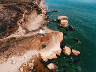 aerial shot of the bride and groom who are standing on the rocks by the ocean