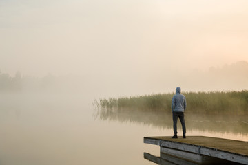 Young man standing alone on wooden footbridge and staring at lake. Mist over water. Foggy air. Early chilly morning. Empty place for sentimental, inspirational text, quote or sayings. Back view. - obrazy, fototapety, plakaty