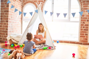 Young beautiful teacher reading book to toddler inside tipi at kindergarten