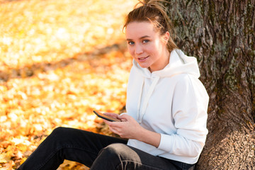Obraz premium Woman in a white sweater with a hood sits on ground in the park near the tree and holding a cellphone in her hands