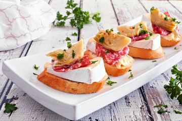 Crostini appetizers with brie cheese, salami and artichokes. Close up on a serving plate against a...