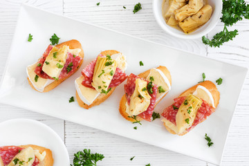 Crostini appetizers with brie cheese, salami and artichokes. Top view on a serving plate against a white wood background. Party food concept.