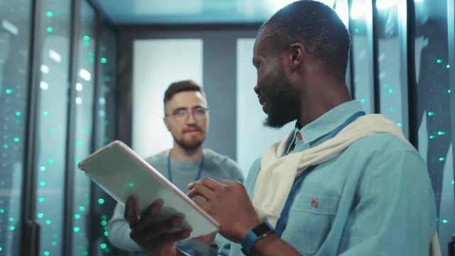 Frustrated upset african administrator noticing data error on digital tablet calling for colleague trying to find solution while coworking in server room.