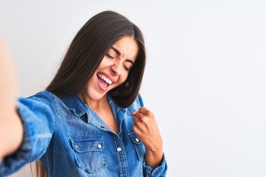 Beautiful woman wearing denim shirt make selfie by camera over isolated white background very happy and excited doing winner gesture with arms raised, smiling and screaming for success