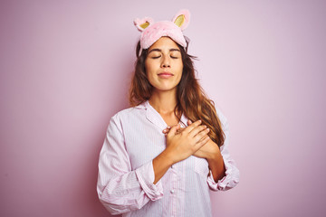 Obraz na płótnie Canvas Young woman wearing pajama and sleep mask standing over pink isolated background smiling with hands on chest with closed eyes and grateful gesture on face. Health concept.