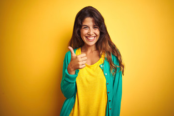 Young woman wearing t-shirt and green sweater standing over yelllow isolated background doing happy thumbs up gesture with hand. Approving expression looking at the camera with showing success.