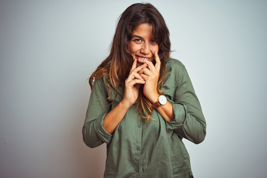 Young beautiful woman wearing green shirt standing over grey isolated background laughing and embarrassed giggle covering mouth with hands, gossip and scandal concept