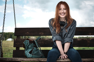 Redhead girl in a denim shirt sits with a backpack on a wooden bench swing widely smiles looking into the frame.