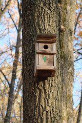 On the trunk of a large tree hangs a wooden birdhouse. Birdhouse on a background of blue sky and trees with yellow leaves. Protection and care of birds in a city park.