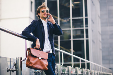 A Nice-Looking Male Boss In Formal Suit And A Briefcase In His Hand Is Smiling While Talking On The Phone In The Street