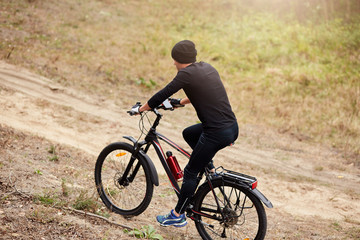 Side wayof male mountain bike cyclist riding uphill along country road, guy wearing black sportwear and cap, sportsman covering assigned distance, enjoying his hobby during vacation. Sport concept.