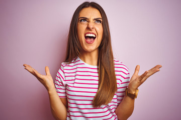 Young beautiful woman wearing striped t-shirt standing over isolated pink background crazy and mad...
