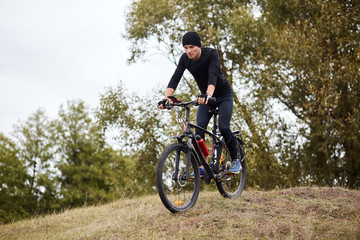 Naklejka na ściany i meble Attaractive man wearing blacktrack suit and cap riding dowhill on his mountain bike, having cardio training, enjoying his recreating in open air and beautiful nature. Sport, healthy lifestyle concept.