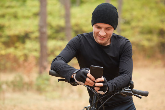 Outdoor shot of young European rider standing in forest and holding his mobile phone, using device for checking social net work while he stops to have rest, enjoyng to ride bike in wood or park.