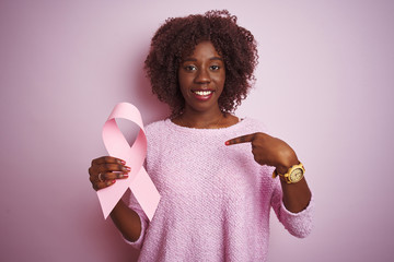 Young african afro woman holding cancer ribbon standing over isolated pink background with surprise...