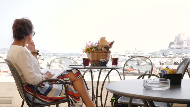 Young woman writing notes on diary at bar table outside. Ponza Island, Italy, sea and cityscape in the background.