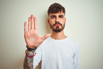 Young man with tattoo wearing t-shirt standing over isolated white background doing stop sing with...