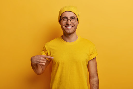 Half length shot of cheerful man points at mockup yellow t shirt, shows space for your design, has glad expression, advertises new outfit, poses against bright background. Look at this t shirt
