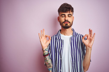 Naklejka premium Young man with tattoo wearing striped shirt standing over isolated pink background relax and smiling with eyes closed doing meditation gesture with fingers. Yoga concept.