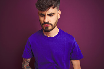 Young man with tattoo wearing t-shirt standing over isolated purple background with hand on stomach because nausea, painful disease feeling unwell. Ache concept.