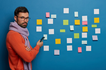 Male student checks work plan, uses modern smartphone, stands in profile, wears glasses, scarf and sweater, prepares for creating presentation, has productive work, stickes glued on blue wall
