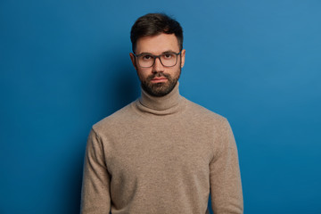 Portrait of serious handsome male has dark hair, thick bristle, looks straightly at camera, wears optical glasses and turtleneck jumper, isolated over blue background, listens attentively interlocutor