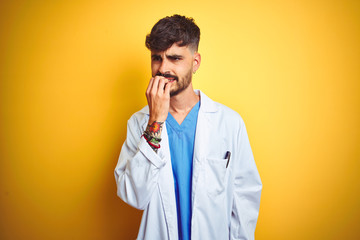 Young doctor man with tattoo standing over isolated yellow background looking stressed and nervous with hands on mouth biting nails. Anxiety problem.