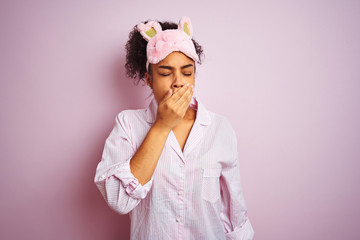 Young african american woman wearing pajama and mask over isolated pink background bored yawning tired covering mouth with hand. Restless and sleepiness.