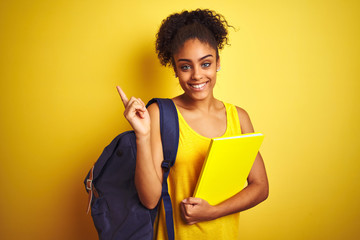 American student woman wearing backpack holding notebook over isolated yellow background very happy...