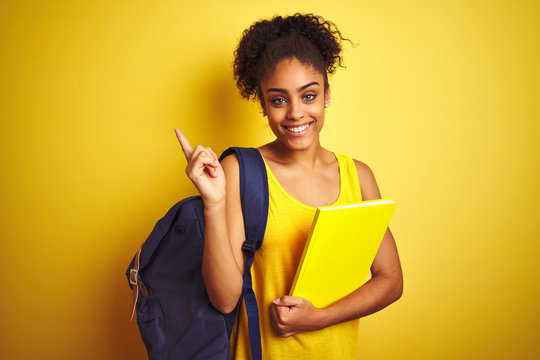 American student woman wearing backpack holding notebook over isolated yellow background very happy pointing with hand and finger to the side