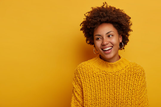 Tender charming happy curly woman has relaxed joyful face expression, Afro hairstyle, wears knitted sweater, laughs enthusiastic, poses against yellow background, blank space aside, turns away