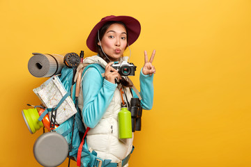 Pretty active backpacker makes victory gesture, keeps lips rounded, holds retro camera, stands with...