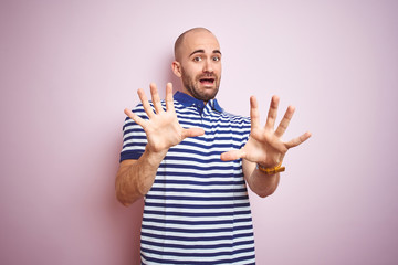 Young bald man with beard wearing casual striped blue t-shirt over pink isolated background afraid and terrified with fear expression stop gesture with hands, shouting in shock. Panic concept.