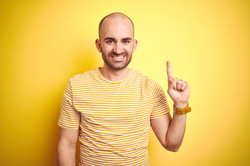 Young bald man with beard wearing casual striped t-shirt over yellow isolated background showing and pointing up with finger number one while smiling confident and happy.