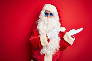 Fototapeta na wymiar Middle age man wearing Santa Claus costume and sunglasses over isolated red background amazed and smiling to the camera while presenting with hand and pointing with finger.