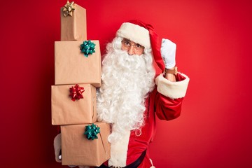 Obraz na płótnie Canvas Senior man wearing Santa Claus costume holding tower of gifts over isolated red background annoyed and frustrated shouting with anger, crazy and yelling with raised hand, anger concept
